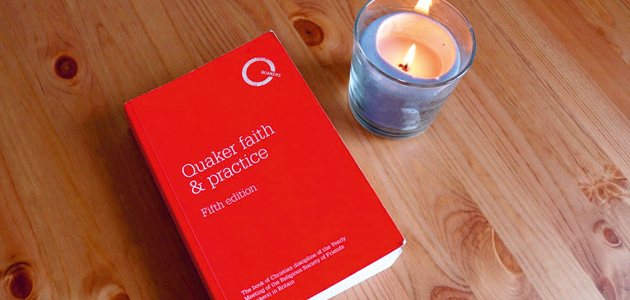 A wooden table top with a book with a red cover and a lit candle